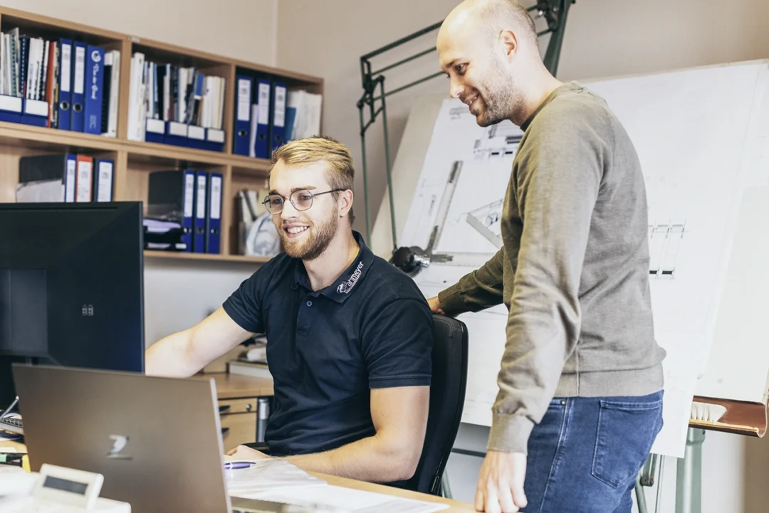 Zwei Mitarbeiter in der technischen Entwicklung besprechen CAD-Konstruktionen am Bildschirm – moderne Büroausstattung mit technischen Zeichnungen im Hintergrund.
